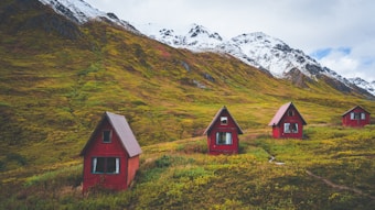 Four small red cabins with triangular roofs are scattered across a grassy hillside. The background features impressive snow-capped mountains under a cloudy sky, suggesting a remote, serene mountain setting.