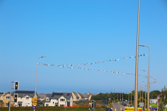 A suburban landscape featuring a clear blue sky and a series of residential buildings with gabled roofs. Colorful triangular flags hang from tall streetlights, adding a festive touch to the scene. Traffic lights and signposts are visible along a road that stretches through the suburb.