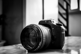 A DSLR camera with a large lens is placed on a reflective surface. The image is in black and white, highlighting the details and textures of the camera body and lens. Background elements are blurred, drawing attention to the camera.