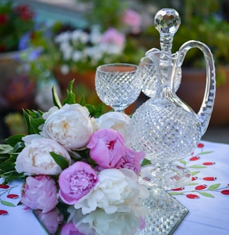 A crystal decanter with a matching glass sits elegantly on a table, accompanied by a bouquet of pink and white peonies. The tablecloth features a pattern of small red and green designs, adding a touch of color. The setting appears to be outdoors, as suggested by the blurred background of greenery and flowers.