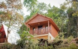 A wooden cabin elevated on a hillside, surrounded by lush greenery and trees. The cabin features a balcony with a simple design and a sloped roof, incorporating rustic charm with its natural materials. Adjacent is a second structure with a similar aesthetic.