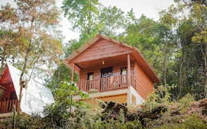 A wooden cabin elevated on a hillside, surrounded by lush greenery and trees. The cabin features a balcony with a simple design and a sloped roof, incorporating rustic charm with its natural materials. Adjacent is a second structure with a similar aesthetic.