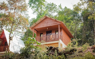 A wooden cabin elevated on a hillside, surrounded by lush greenery and trees. The cabin features a balcony with a simple design and a sloped roof, incorporating rustic charm with its natural materials. Adjacent is a second structure with a similar aesthetic.