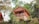 A wooden cabin elevated on a hillside, surrounded by lush greenery and trees. The cabin features a balcony with a simple design and a sloped roof, incorporating rustic charm with its natural materials. Adjacent is a second structure with a similar aesthetic.