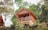 A wooden cabin elevated on a hillside, surrounded by lush greenery and trees. The cabin features a balcony with a simple design and a sloped roof, incorporating rustic charm with its natural materials. Adjacent is a second structure with a similar aesthetic.
