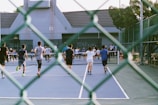 A group of people playing or participating in an activity on a tennis court. The view is partially obscured by a chain-link fence in the foreground. The participants, mostly wearing shorts and T-shirts, are actively moving towards the net. The background includes a building with a gabled roof and surrounding trees, adding to the setting of an outdoor sports facility.