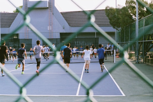 A group of people playing or participating in an activity on a tennis court. The view is partially obscured by a chain-link fence in the foreground. The participants, mostly wearing shorts and T-shirts, are actively moving towards the net. The background includes a building with a gabled roof and surrounding trees, adding to the setting of an outdoor sports facility.