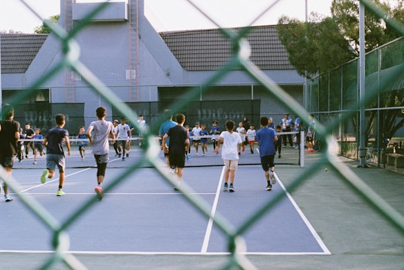 A group of people playing or participating in an activity on a tennis court. The view is partially obscured by a chain-link fence in the foreground. The participants, mostly wearing shorts and T-shirts, are actively moving towards the net. The background includes a building with a gabled roof and surrounding trees, adding to the setting of an outdoor sports facility.