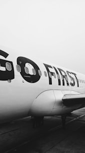 An airplane with the words 'GO FIRST' printed in large letters on the side. The image is in black and white, emphasizing the airplane's sleek design and the text. The foreground shows part of the wing and engines, while the background is a cloudy sky.