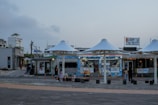 A coastal commercial area featuring open stalls with large white umbrellas, surrounded by various shops. Several people are visible, including children, engaged in casual activities near the structures. There's signage with text in another language and visible electrical lines in the background.