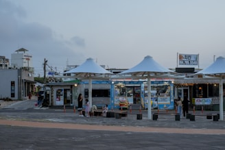 A coastal commercial area featuring open stalls with large white umbrellas, surrounded by various shops. Several people are visible, including children, engaged in casual activities near the structures. There's signage with text in another language and visible electrical lines in the background.