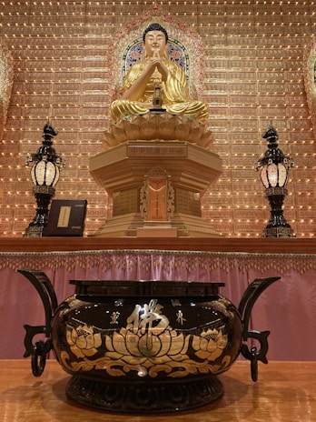A golden Buddha statue sits prominently on an ornate pedestal, surrounded by detailed wall carvings and a backdrop filled with small illuminated spaces. Flanking the statue are two intricately designed lamps. In the foreground, a large black and gold incense burner with floral patterns is placed on a polished wooden surface.
