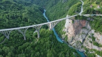 A picturesque bridge stretches across a deep, green forested canyon with a turquoise river flowing beneath. The bridge has elegant arches and is surrounded by lush greenery and rocky cliffs. A winding road leads to a small structure nestled amongst the trees.