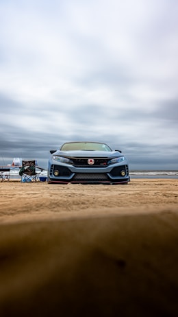 A silver car with a red logo sits on the beach sand, flanked by two folding chairs. The backdrop is an overcast sky with rolling clouds above a vast stretch of shoreline.