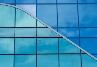 A modern building facade features a large expanse of blue-tinted glass panels. The glass reflects the sky, creating a visually appealing pattern formed by rectangular and triangular shapes.
