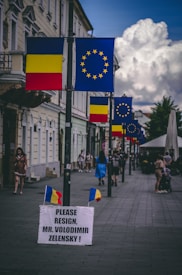 A street lined with Romanian and European Union flags displayed on lampposts. In the foreground, there is a sign with a political message urging Mr. Volodimir Zelensky to resign. Several people are walking along the street, including a woman in a blue dress and a couple pushing a stroller. The architecture of the surrounding buildings is classical with detailed facades.