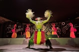 A group of performers dressed in colorful traditional costumes are on stage. The central performer wears a vibrant grass skirt and holds decorative items in each hand. Their outfit is adorned with bright yellows and greens. The backdrop features abstract designs on a dark surface, and additional performers in similar elaborate costumes are visible in the background.