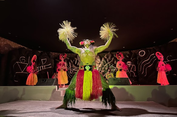 A group of performers dressed in colorful traditional costumes are on stage. The central performer wears a vibrant grass skirt and holds decorative items in each hand. Their outfit is adorned with bright yellows and greens. The backdrop features abstract designs on a dark surface, and additional performers in similar elaborate costumes are visible in the background.
