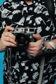 Hands holding a vintage camera with a black and white patterned shirt in the background. The shirt features abstract and floral designs, and there is a blue background visible.
