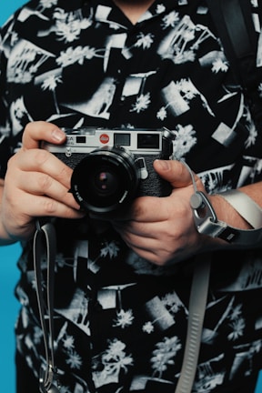 Hands holding a vintage camera with a black and white patterned shirt in the background. The shirt features abstract and floral designs, and there is a blue background visible.