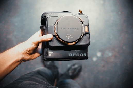 A hand holds a square-shaped electronic device with a prominent circular dial in the center, branded 'PolarPro', against a blurred background. The device appears to be a camera filter or lens system.