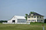 A large farmhouse with a yellow facade and black shutters sits next to a white barn. The structures are surrounded by a neatly trimmed lawn and fenced by a low white fence. The sky is clear, and there are a few scattered trees in the background.