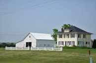 A large farmhouse with a yellow facade and black shutters sits next to a white barn. The structures are surrounded by a neatly trimmed lawn and fenced by a low white fence. The sky is clear, and there are a few scattered trees in the background.