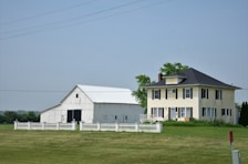 A large farmhouse with a yellow facade and black shutters sits next to a white barn. The structures are surrounded by a neatly trimmed lawn and fenced by a low white fence. The sky is clear, and there are a few scattered trees in the background.