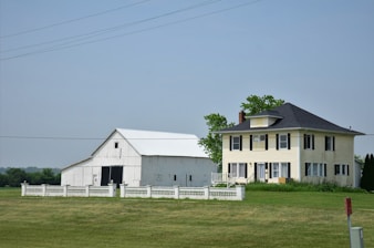 A large farmhouse with a yellow facade and black shutters sits next to a white barn. The structures are surrounded by a neatly trimmed lawn and fenced by a low white fence. The sky is clear, and there are a few scattered trees in the background.