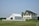 A large farmhouse with a yellow facade and black shutters sits next to a white barn. The structures are surrounded by a neatly trimmed lawn and fenced by a low white fence. The sky is clear, and there are a few scattered trees in the background.