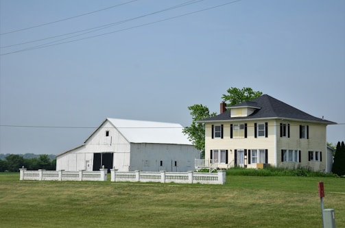 A large farmhouse with a yellow facade and black shutters sits next to a white barn. The structures are surrounded by a neatly trimmed lawn and fenced by a low white fence. The sky is clear, and there are a few scattered trees in the background.