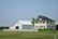 A large farmhouse with a yellow facade and black shutters sits next to a white barn. The structures are surrounded by a neatly trimmed lawn and fenced by a low white fence. The sky is clear, and there are a few scattered trees in the background.