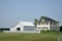 A large farmhouse with a yellow facade and black shutters sits next to a white barn. The structures are surrounded by a neatly trimmed lawn and fenced by a low white fence. The sky is clear, and there are a few scattered trees in the background.