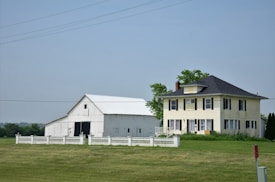 A large farmhouse with a yellow facade and black shutters sits next to a white barn. The structures are surrounded by a neatly trimmed lawn and fenced by a low white fence. The sky is clear, and there are a few scattered trees in the background.