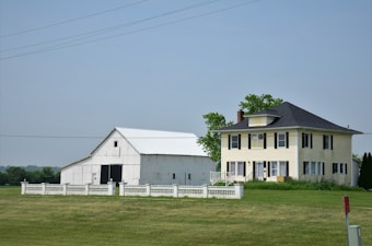 A large farmhouse with a yellow facade and black shutters sits next to a white barn. The structures are surrounded by a neatly trimmed lawn and fenced by a low white fence. The sky is clear, and there are a few scattered trees in the background.