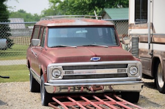 A vintage Chevrolet vehicle with a rusty, faded red exterior is parked on a gravel path. The car appears weathered, with visible signs of age, surrounded by a chain-link fence and other vehicles in a yard. The background includes some green foliage and a building with a green roof.