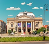 A grand, historic theater building with ornate architectural features and columns stands prominently in a plaza. In front of the theater, a statue is situated on a pedestal amid landscaped greenery. Banners hang on the building's facade, and two parked cars are visible on the side. The sky above is blue with scattered fluffy clouds, and a vintage-style street lamp is present near the foreground.