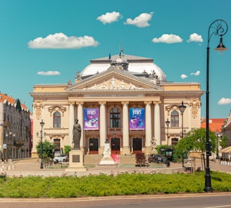 A grand, historic theater building with ornate architectural features and columns stands prominently in a plaza. In front of the theater, a statue is situated on a pedestal amid landscaped greenery. Banners hang on the building's facade, and two parked cars are visible on the side. The sky above is blue with scattered fluffy clouds, and a vintage-style street lamp is present near the foreground.