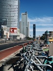 A row of rental bicycles is lined up along a bustling city street with several high-rise buildings. The sky is clear and blue, and there is a red painted bike lane adjacent to the sidewalk. Advertisements are visible on the bikes and nearby billboards.