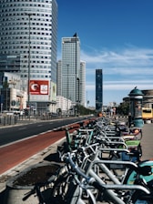 A row of rental bicycles is lined up along a bustling city street with several high-rise buildings. The sky is clear and blue, and there is a red painted bike lane adjacent to the sidewalk. Advertisements are visible on the bikes and nearby billboards.