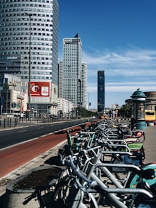 A row of rental bicycles is lined up along a bustling city street with several high-rise buildings. The sky is clear and blue, and there is a red painted bike lane adjacent to the sidewalk. Advertisements are visible on the bikes and nearby billboards.