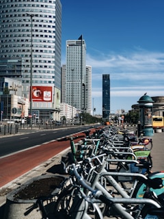A row of rental bicycles is lined up along a bustling city street with several high-rise buildings. The sky is clear and blue, and there is a red painted bike lane adjacent to the sidewalk. Advertisements are visible on the bikes and nearby billboards.
