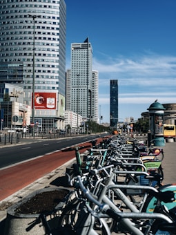 A row of rental bicycles is lined up along a bustling city street with several high-rise buildings. The sky is clear and blue, and there is a red painted bike lane adjacent to the sidewalk. Advertisements are visible on the bikes and nearby billboards.
