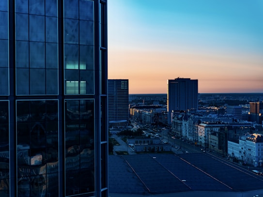 A cityscape displays tall modern buildings with reflective glass windows on the left. As the sun sets, the sky transitions from a light orange near the horizon to a deep blue at the top. Urban streets and smaller buildings can be seen extending into the distance.