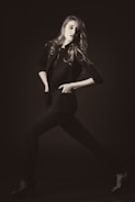 A stylishly dressed individual poses dynamically in a monochrome setting, exuding confidence. The person wears a tailored jacket and heels, with hair flowing elegantly.