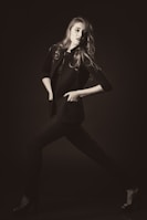 A stylishly dressed individual poses dynamically in a monochrome setting, exuding confidence. The person wears a tailored jacket and heels, with hair flowing elegantly.