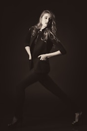 A stylishly dressed individual poses dynamically in a monochrome setting, exuding confidence. The person wears a tailored jacket and heels, with hair flowing elegantly.