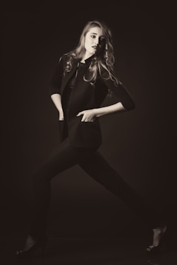 A stylishly dressed individual poses dynamically in a monochrome setting, exuding confidence. The person wears a tailored jacket and heels, with hair flowing elegantly.