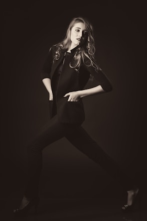 A stylishly dressed individual poses dynamically in a monochrome setting, exuding confidence. The person wears a tailored jacket and heels, with hair flowing elegantly.