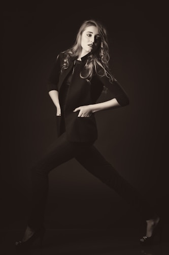 A stylishly dressed individual poses dynamically in a monochrome setting, exuding confidence. The person wears a tailored jacket and heels, with hair flowing elegantly.